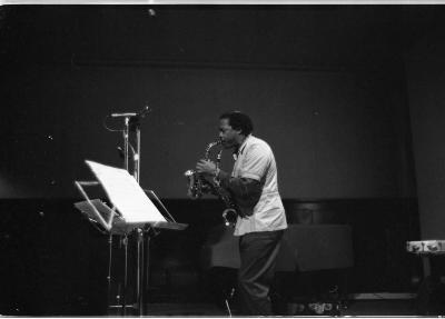 A side profile long-shot musician wearing a light coloured shirt and dark pants as he plays the saxophone. In front of him are three microphones resting on stands, and in the foreground there’s a music stand with sheet music.