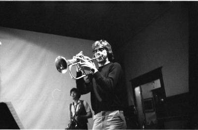 A low angle shot of a trumpet player. He’s wearing a dark shirt, jeans and aviator sunglasses. A man playing the saxophone is behind him. The background contains a large white tarp, and a doorframe. The image is in black and white.