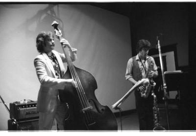 There are two men playing instruments. The man on the left wears a suit and he is playing a cello. The man on the right wears a vest, button-up shirt, and dark pants as he plays the saxophone. Each musician has a music stand in front of him, and several pieces of other equipment behind them. The image is in black and white.
