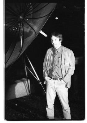 A dark-haired man poses with his hands in his pockets next to a large satellite dish, presumably on a rooftop. He wears a dark dress shirt tucked into white trousers, a light-colored blazer and a plaid tie. 
