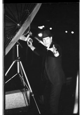 A man in a dark suit and a fedora stands beside a rooftop satellite dish, illuminated by a bright camera flash. His right hand is positioned on the dish while the left reaches out towards the camera, his expression somewhere between cheeky and scandalized. 
