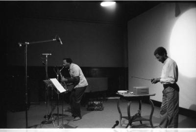 A long-shot of a man playing the saxophone as he bends closer to the microphone in front of him. To his side there are two other microphones and a music stand with sheet music. Another man stands behind him, next to a table, as he holds a violin bow in his hands.