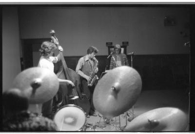 A high-angle shot behind the drummer featuring three cymbals and parts of the drum-set. Other bandmates are featured behind the drumset, a cello, saxophone, and trumpet player. The image is in black and white.