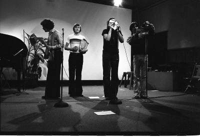 Four people stand before three stand up microphones, all engaged in a different activity. One person is looking to their right, inquisitive, holding a notebook. Another stands front facing with a blank expression, holding a brown paper bag and a drink. A third sticks their tongue through a ripped sheet of paper. The fourth is decanting liquid into a glass in front of the microphone, as though to capture the sound. 