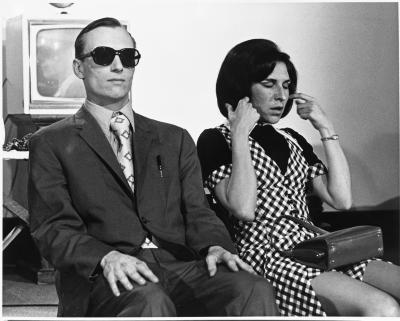 Randy Gledhill and Berenicci Hershorn sit side by side on the set of As the World Burns. Randy wears a suit and patterned tie, and sunglasses. Wearing a patterned mini dress, Berenicci balances a purse on her lap. She is caught mid-blink while tucking her bob behind her ears. 