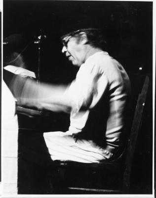 A high contrast image of a man energetically playing piano. He is seen in profile and wears light coloured clothing, glasses. His hands and arms are blurred as he presses the piano keys. This image is in black and white.