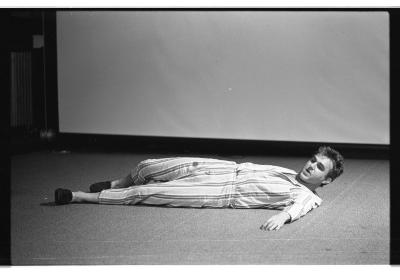 A man is lying down on the floor, with his left arm extended out on the floor and his right knee propped up, as he cranes his head up looking to the left side of the image. His hair is barbered and he is wearing a matching striped shirt and pants with slippers. In the background, there is a projector screen and to the left of it is a radiator. The image is in black and white.
