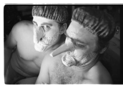 A black-and-white portrait of Martin von Haselberg and Brian Routh. They are both shirtless, and are wearing shower caps and prosthetic noses and chins. Their faces are painted white with black dots to create the effect of stubble. 