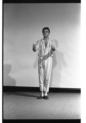 A man standing with his arms extended forward. His hands are blurred and both his index fingers point out. His hair is barbered, he is wearing a matching striped shirt and pants with slippers. His gaze looks off to the left of the image. In the background there is a projector screen. The image is in black and white.
