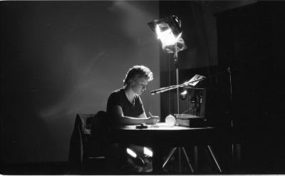 A large stage light stands tall, and is pointed down toward a person sitting at a desk with four small objects. They have short feathered hair, and hold a pen in their right hand and address the surface below them. A microphone protrudes from the right side and is set close to the person’s mouth. 
