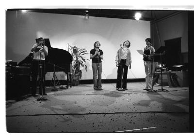 Four people perform passionately in front of four stand up microphones. One performer is playing the trumpet. The ground is littered with debris and wires from the microphones. They stand in front of a piano, a plant, and a blank projection screen. 