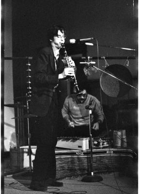 A man plays the clarinet, he is wearing a dark coloured suit and glasses. Two microphones on stands are positioned in front of him, and behind him there is a gong suspended in air and a musician is seated playing several instruments. 