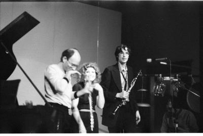 A musician laughs into his hand as he holds his flute with the other, the woman next to him looks towards him and laughs as well. While a man wearing a dark suit and glasses holds a clarinet in his hand as he looks out. To the front and side of the group there is sound equipment and a man crouched behind it. The corner shows part of a grand piano. 