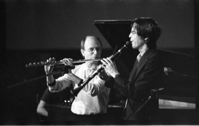 Two musicians play the flute and clarinet. The clarinettist wears a dark blazer and glasses while the flautist standing behind him wears a long-sleeve t-shirt and glasses. Behind the musicians someone plays the grand piano.