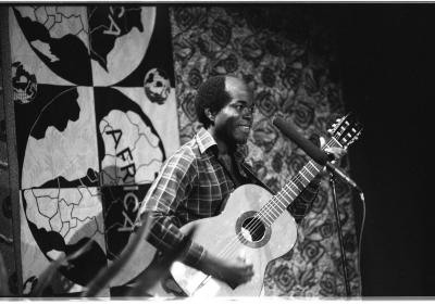 Jacob Pereira-Lunghu smiles towards the audience while playing acoustic guitar. He wears a plaid button-up shirt and performs in front of a patterned backdrop with a stylized map of Africa. The photo is in black-and-white. 