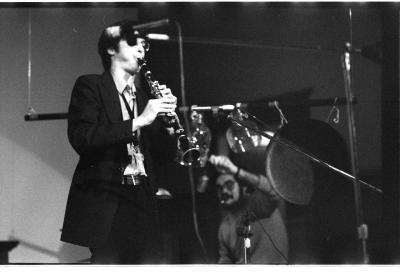 A clarinettist performs, he's wearing a dark suit and glasses. Two microphones are on stands in the foreground, and a man holding a microphone records him in the background.