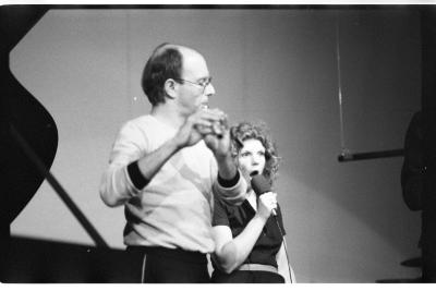 A flautist performs with a woman as she sings into a wired microphone. The corner of the image depicts part of a grand piano.