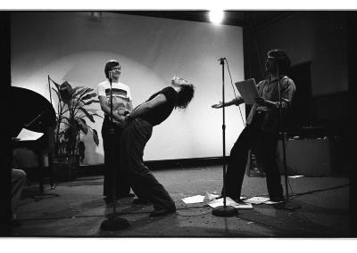 Three people perform in front of three stand up microphones. On the right, a performer is giving an operatic belt with one hand outstretched and a notebook in the other. Centrally, a performer leans back with one hand to their groin and a drink in the other. The third performer stands a few meters back, holding a brown paper  bag with both hands and wearing a grin. 