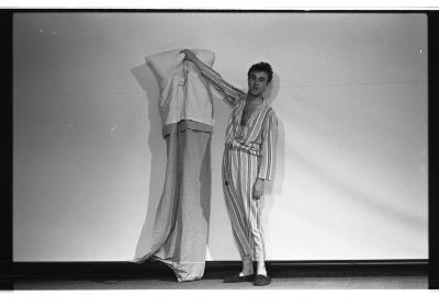 A man is standing as he holds a sleeping bag up high with his right arm. His hair is barbered, he is wearing a matching striped shirt and pants with slippers. His gaze looks off to the left of the image. In the background there is a projector screen. The image is in black and white

