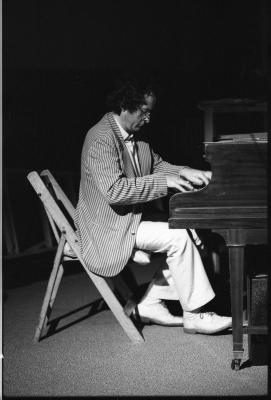 Side profile of a dark-haired man sitting on a folding chair, looking down at his hands as he plays the piano. He wears glasses, a pinstripe blazer over a white dress shirt, white trousers and white dress shoes.
