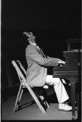 Side profile of a dark-haired man sitting on a folding chair, his head tilted backwards with great dramatism as he plays the piano. He wears glasses, a pinstripe blazer, white trousers, and white dress shoes. 
