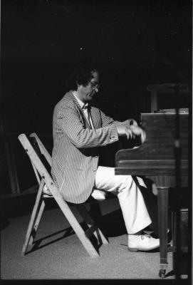 Side-profile of a dark-haired man sitting on a folding chair, looking down as he plays the piano. He is caught mid-action, his hands blurred with movement. He wears glasses, a pinstripe blazer, white trousers and white dress shoes. 
