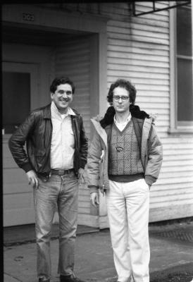 Two dark-haired men pose in front of the Western Front, one smiling and one somber. The smiling man wears a leather jacket over a light-colored dress shirt tucked into dark-wash jeans. The somber man wears glasses, a puffer jacket and a patterned vest over a white dress shirt and white trousers. 
