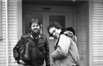 A bearded man stands in front of the doors of the Western Front, smiling at the camera with kind eyes. He wears a leather jacket, a dark vest, and a dress shirt. He holds a messenger bag under his right arm. To his left a man with disheveled curls and glasses stands with his side to the camera, smiling slyly with his head tilted. He wears a fur-lined puffer jacket. 
