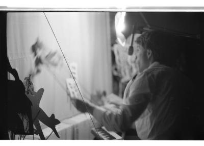 A puppeteer backstage controls shadow puppets along the portable shadow theatre. A lamp shines on the theatre screen. 
