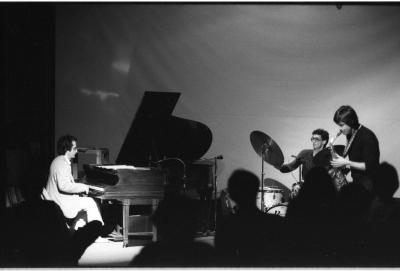 We peer over the back of silhouetted audience members’ heads as a band plays on stage at the Grand Luxe Hall. To the left, a dark-haired man in a light-colored suit plays the piano. Beside him is a dark-haired drummer sitting behind a white drum kit. In front of the drummer is a saxophonist dressed in all black, leaning forward slightly as he plays. 
