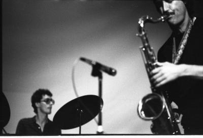 Quarter-profile view of a saxophonist from eye to hip fills the left portion of the frame. His dark mustache matches his all-black attire, his instrument glinting as he plays with deep focus. In the middleground stands a blurry microphone, pointing left. In front of the microphone is the silhouette of two cymbals, resembling black discs. Behind them is the small figure of a drummer, captured chest-up, as he looks to the side. He has dark curls and wears aviator glasses and a black shirt. 
