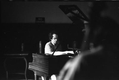 We peer over a blurry audience member’s shoulder at a dark-haired man playing the piano in the Grand Luxe Hall. He wears glasses and a pinstriped top, peering down at his sheet music with deep focus. Behind him, a small sign on the wall reads NO SMOKING.
