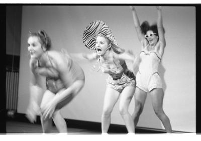 Three unorthodox women in 50s-style bathing suits stand in a line. At the front, a woman in pigtails jumps, her entire body blurred by movement. In the middle, a woman in a floral one-piece and a zebra-print sun hat screams, her arms blurring as she waves them. At the back, a dark-haired woman wearing white sunglasses holds her hands up in the air.
