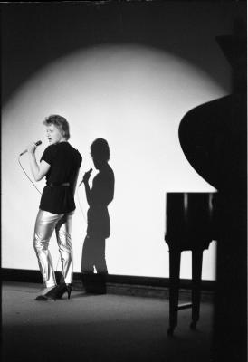 A large spotlight creates a crescent shape across a flat surface and highlights a person wearing kitten heels and shiny pants who stands in front of the smooth background. They hold a wired microphone in their left hand to their mouth, and their silhouette is mirrored behind them. 
