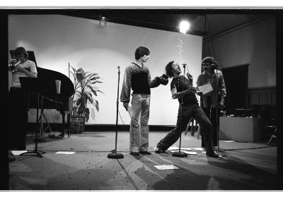 Three performers stand in front of three stand up microphones. Debris litters around their feet. Liquid hangs in mid-air. The performer in the middle of the three attempts to catch it in their mouth. The performer to the right stands idly with a jug and the performer to the left wears a concerned expression. There is a fourth performer on the far left side of the image playing the trumpet. 