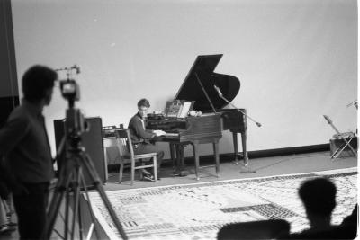 David Weinstein plays the piano in the Grand Luxe Hall. He twists in his chair to read notation on a room-sized graphic score laid out on the floor. A camera person documents the performance from behind a tripod. The image is in black and white.