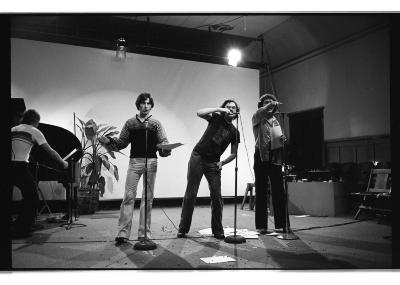 Three people perform in front of stand up microphones. On the left a person stands with their arms outstretched and shrugged, the middle performer gestures as if putting food into their mouth, and on the right the performer points out in front of them with their left arm. They all stand in front of a blank projection screen. 