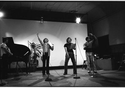 Four performers stand before stand up microphones all in different phases of expression. One watches intently, holding a brown paper bag with both arms. The other three performers flail an arm in passionate expression. 