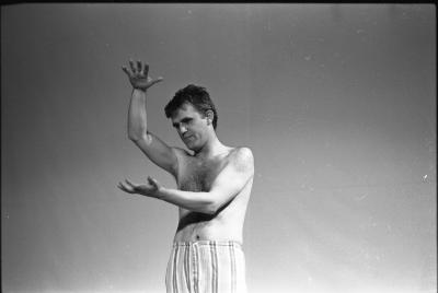 A medium-long shot of a man standing creating gestures with his arms and hands. His hair is barbered, he is shirtless and wearing striped pants with slippers. In the background there is a projector screen. The image is in black and white.
