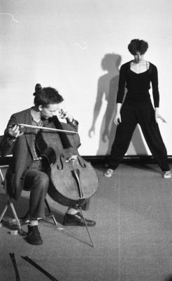 Ernst Reijseger sits on a chair while playing cello. Behind him, Jennifer Mascall stands in a wide legged posture with her arms by her sides. The image is in black and white.