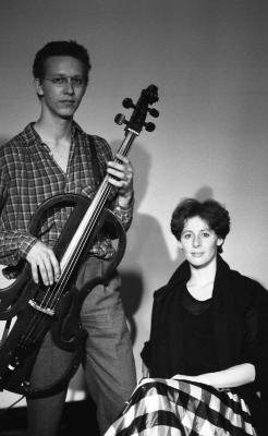 Wearing glasses and a plaid shirt, Ernst Reijseger stands holding a cello with a cutout frame while Jennifer Mascall sits next to him, smiling at the camera. 