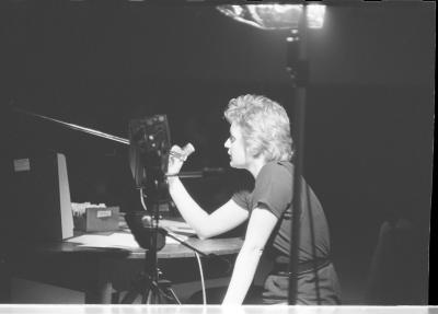  A person is highlighted by a standing stage light that emphasises their body, and sets them in direct contrast to the space they are situated in. A series of paper objects are scattered on the desk in front of them. The person rests their left hand on their thigh, and their right elbow is propped on the desk. 
