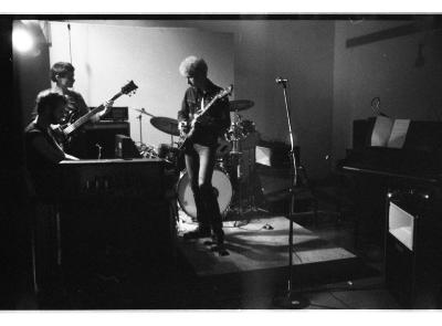 Three people play music in a band room setup. Two people play guitar while one sits to play the piano. 