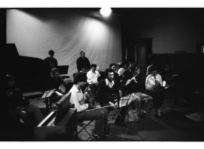A group of thirteen musicians with various string, percussion, and woodwind instruments sit in front of music stands in the Grand Luxe Hall. The photo is in black and white.
