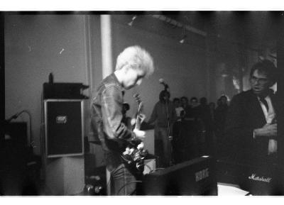 Photographed in black and white. A person with blonde hair wearing a denim jacket and tight denim jeans is playing the guitar to a crowd of people. They are surrounded by musical gear.
