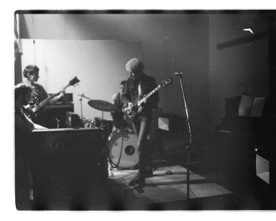 A black and white image. A four piece band plays music together. Two have guitars, one person is sitting at the drum kit, and a fourth member seems to be sitting behind a keyboard. One band member is wearing a black denim jacket and has blonde hair. 