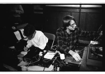 Two people sit side by side at a table covered with music equipment and cables, focused on their respective tasks. One person in a white dress shirt gazes down while writing on a piece of paper. The other person is turning a knob on a synth.