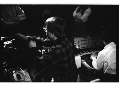 A performer wearing a plaid shirt and glasses is seen from behind as they place a vinyl on a turntable. Next to them, another performer sits in front of a synthesizer with a keyboard. Audience members stand in front of the performers with their arms crossed, but their heads are cropped out of the image.