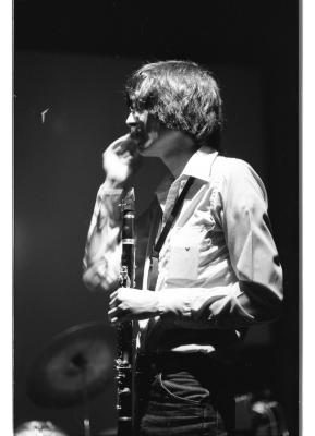 Artist Paul Cram stands holding a clarinet while fixing his moustache. He is on a stage, with a drum kit behind him and lights flooding from the top of the image. 