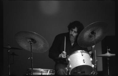 Gregg Simpson sits behind a drumkit with one drumstick raised, ready to play. He has a moustache and wears a black button down shirt. 